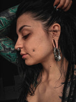 A person with dark hair and various piercings, including a unisex Kaya Movement Darma Biomechanical Clicker Hoop Septum Nose Ring in Silver (16 gauge), ear gauges, and a dermal piercing on the cheek, poses with one arm raised, showcasing their profile. They have tattoos visible on their arm. The background is dark and out of focus.