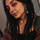 A woman with long, dark dreadlocks and noticeable earrings, including a large spiral gauge, poses for a selfie. Donning post-apocalyptic body jewelry and the Darma Biomechanical Clicker Hoop Septum Nose Ring in Silver by Kaya Movement, she showcases multiple facial piercings: a unisex 16 gauge nose piercing, lip piercing, and a dermal on her cheek. She wears a brown jacket and black top.