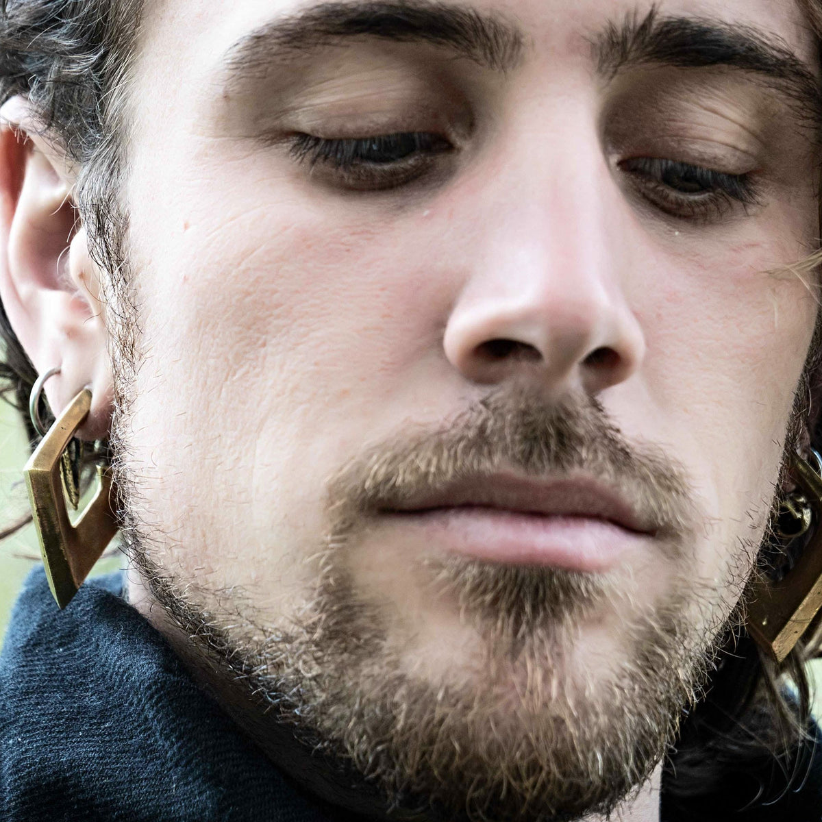 Close-up of a person with a light beard and mustache, wearing large, golden lightning bolt-shaped earrings. The minimalist SAN Minimalist Diamond Ear Weights in Gold by Kaya Movement add an extra touch of elegance. The person has a contemplative expression and is looking down, with the blurred background emphasizing the face and earrings.