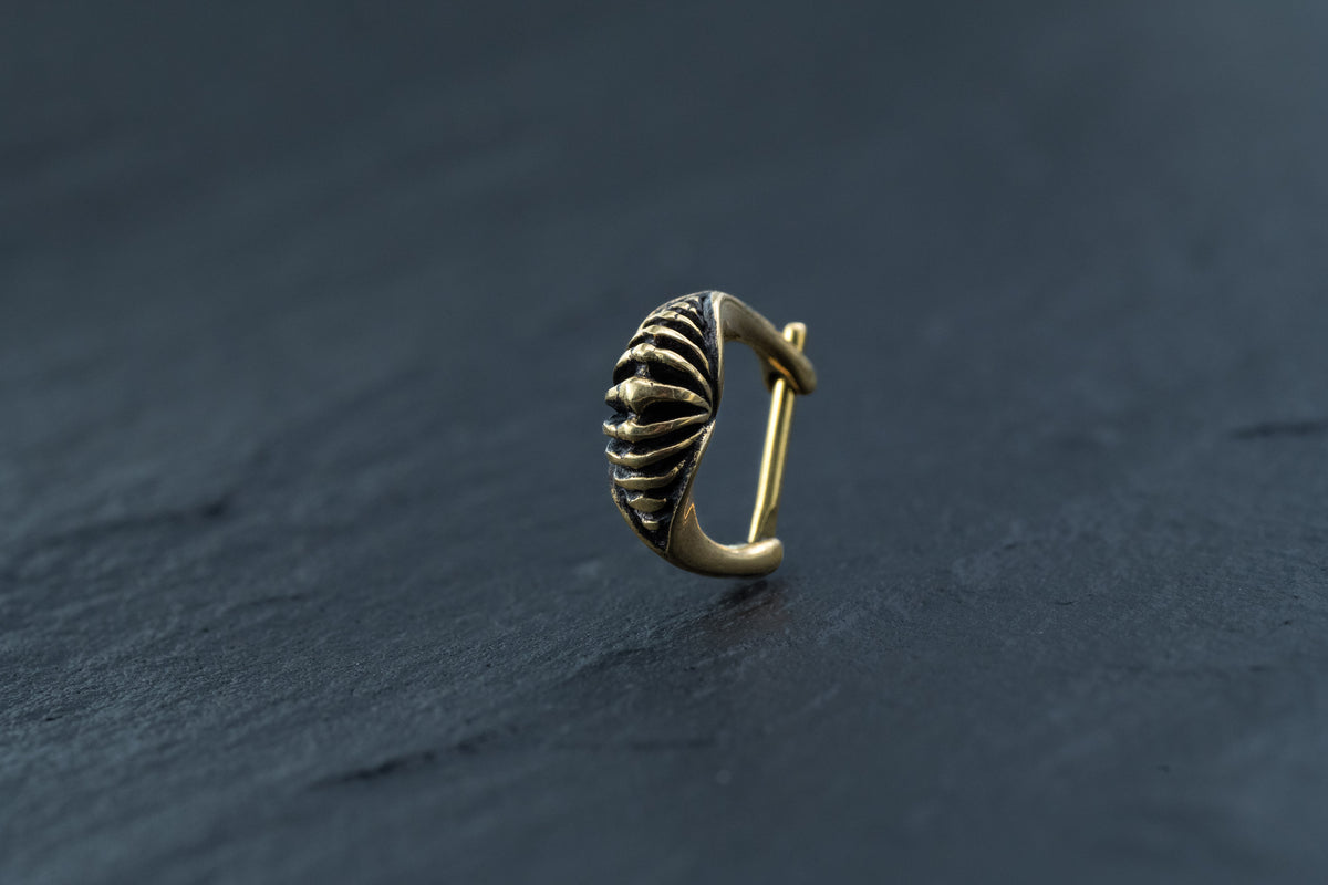 The DIARA Biomechanical Clicker Hoop Septum Nose Ring in Gold by Kaya Movement, featuring an elegant 16-gauge curved hoop with intricate shell-like design and detailed textures, is placed on a dark, smooth surface. This exquisitely crafted nose ring makes an exceptional addition to any unisex piercing collection.