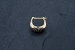 A close-up of the DIARA Biomechanical Clicker Hoop Septum Nose Ring in Gold by Kaya Movement, placed on a dark, textured surface. The hoop earring boasts a unique U-shaped design with a ridged, fan-like accent at its base, reminiscent of a chic unisex nose piercing.