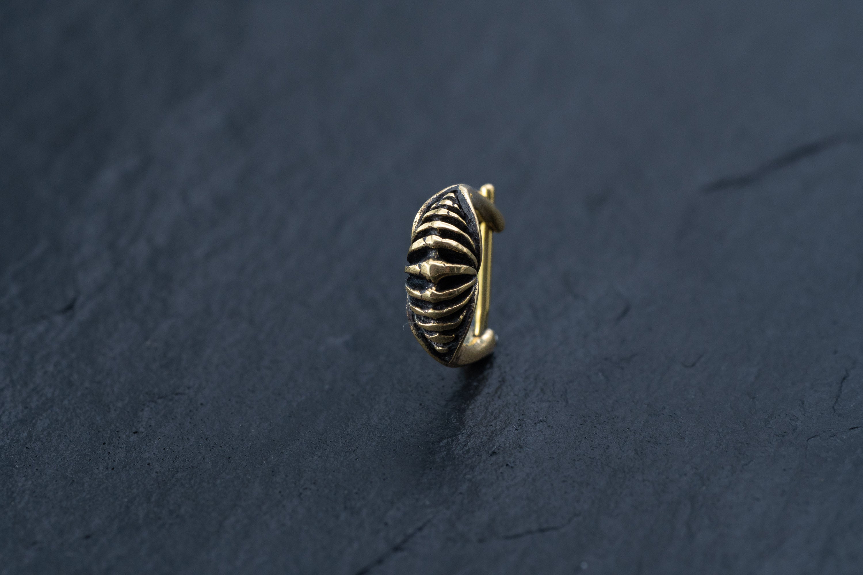 A close-up photograph of the DIARA Biomechanical Clicker Hoop Septum Nose Ring in Gold by Kaya Movement resting on a dark, textured surface. The 16-gauge ring features an intricate, ribbed design that gives it a distinctive, sculptural appearance. The lighting highlights the metallic finish and details of the design.