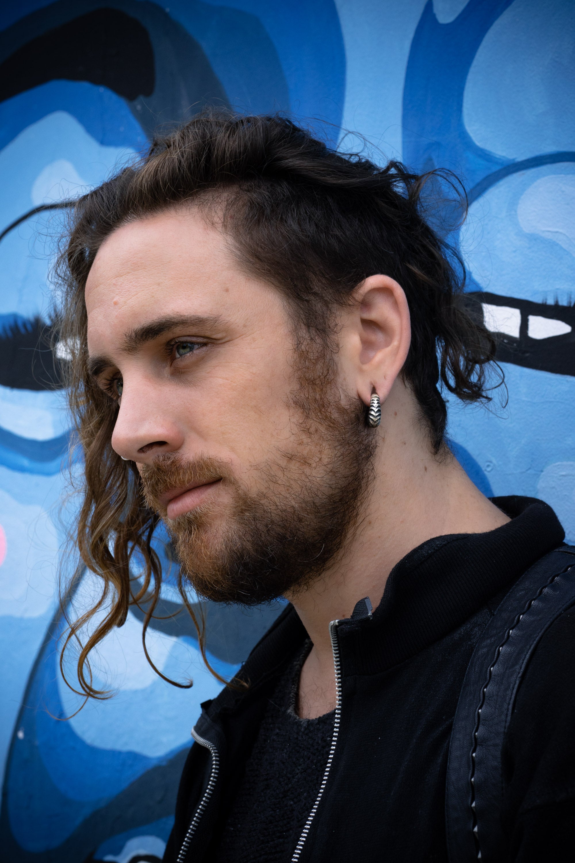 A person with wavy brown hair, a beard, and a septum piercing is looking to the side. They are wearing the AARO Cyborg Clicker Hoop Ear or Septum Weights in 925 Silver by Kaya Movement as both an earring and septum ring. Standing in front of a blue mural featuring abstract shapes and clad in a black jacket, they appear contemplative.