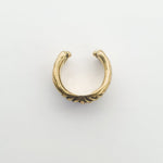 The DARMA Fake Septum Ring or Ear Cuff in Gold by Kaya Movement is showcased against a plain white backdrop. This small, gold-colored piece features an open-ended design and is adorned with a delicate, intricate floral pattern in the center, adding a touch of elegance to its simple, curved form. Its versatility ensures it fits seamlessly into any unisex nose jewelry collection.