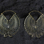 A pair of intricately designed CIRCUIT Oversized Visionary Disc Hoop Earrings in Gold by Kaya Movement lies on a dark textured surface. These tribal earrings feature an elaborate, maze-like pattern with a symmetrical, geometric motif, resembling a stylized bird with outstretched wings at the center of each earring.