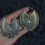 A hand holds Kaya Movement's CIRCUIT Oversized Visionary Disc Hoop Earrings in Gold (16 gauge), featuring a large, intricate, maze-like design and a golden finish. The dark, textured background enhances these boho statement earrings. Their hoop style showcases detailed patterns converging at the center.