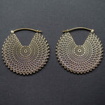 A pair of VORTEX Tribal Oversized Mandala Disc Hoop Earrings in gold, by Kaya Movement, showcasing an intricate latticework with concentric circles and a scalloped edge design, reminiscent of boho statement earrings, laying against a dark gray textured background.