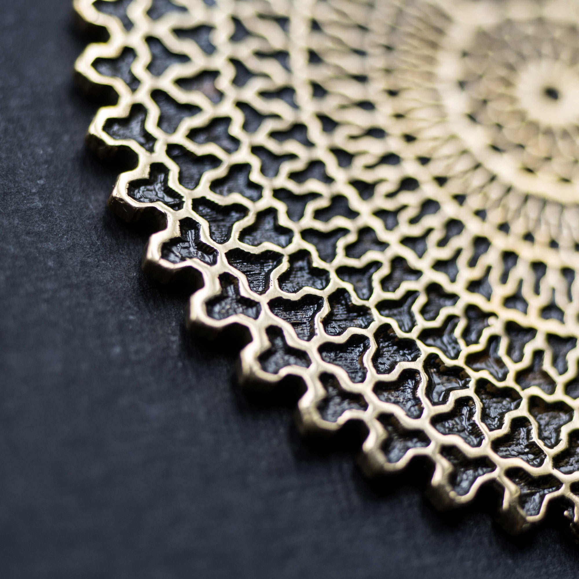 Close-up of the Kaya Movement's VORTEX Tribal Oversized Mandala Disc Hoop Earrings in Gold against a dark gray background. The 16 gauge earrings feature interlocking shapes, forming a detailed and symmetrical mesh with a lace-like appearance. The lighting accentuates the shine and intricate texture of the metallic design.
