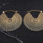A pair of VORTEX Tribal Oversized Mandala Disc Hoop Earrings in Gold by Kaya Movement is displayed on a dark, textured background. These 16-gauge earrings feature a web-like structure with repeating symmetrical shapes radiating from the center.