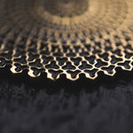 Close-up photo of an intricate gold pattern against a dark background. The design features interconnected geometric shapes, reminiscent of the VORTEX Tribal Oversized Mandala Disc Hoop Earrings in Gold by Kaya Movement, creating a sense of depth and texture. The image is blurred towards the top, drawing focus to the detailed craftsmanship.