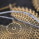Close-up of intricate, gold-colored filigree resembling geometric patterns on a dark background, much like the detailed design found in Kaya Movement's VORTEX Tribal Oversized Mandala Disc Hoop Earrings in Gold. Twigs are placed across the filigree, adding texture and depth to the image.