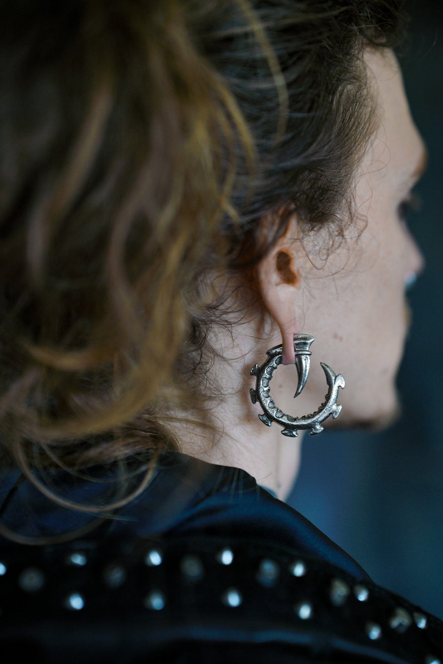 A person with long brown hair tied back is wearing a large, ornate GIGER Biomechanical Open Ended Hoop Ear Weight in Silver by Kaya Movement, featuring a crescent shape with decorative spikes that evoke post-apocalyptic body jewelry. They are also wearing a studded black garment, partially visible, against a blurred background.