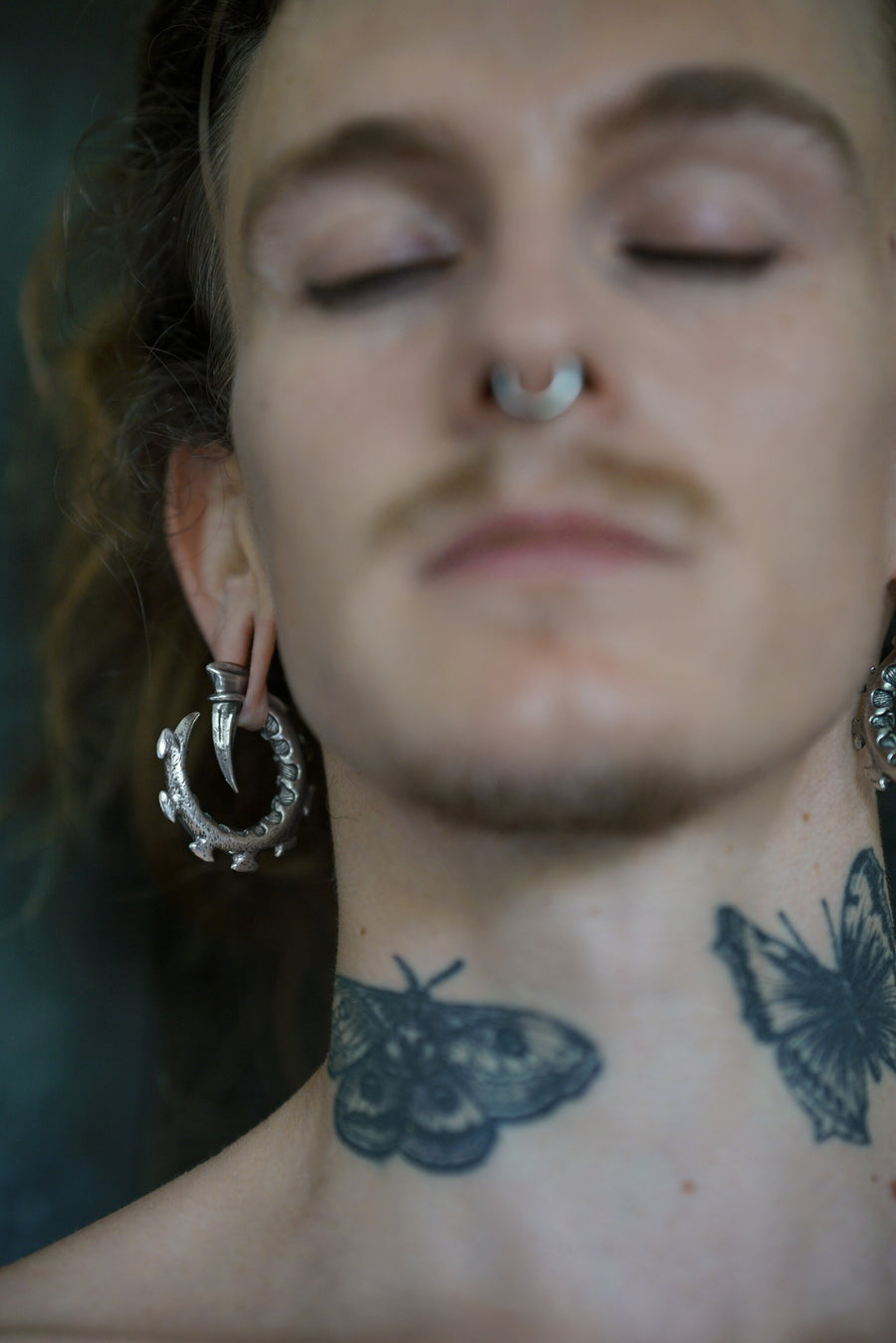 A person with their eyes closed wears a septum nose ring and GIGER Biomechanical Open Ended Hoop Ear Weights in Silver by Kaya Movement. They have visible neck tattoos of a moth and a butterfly. The background is blurred.