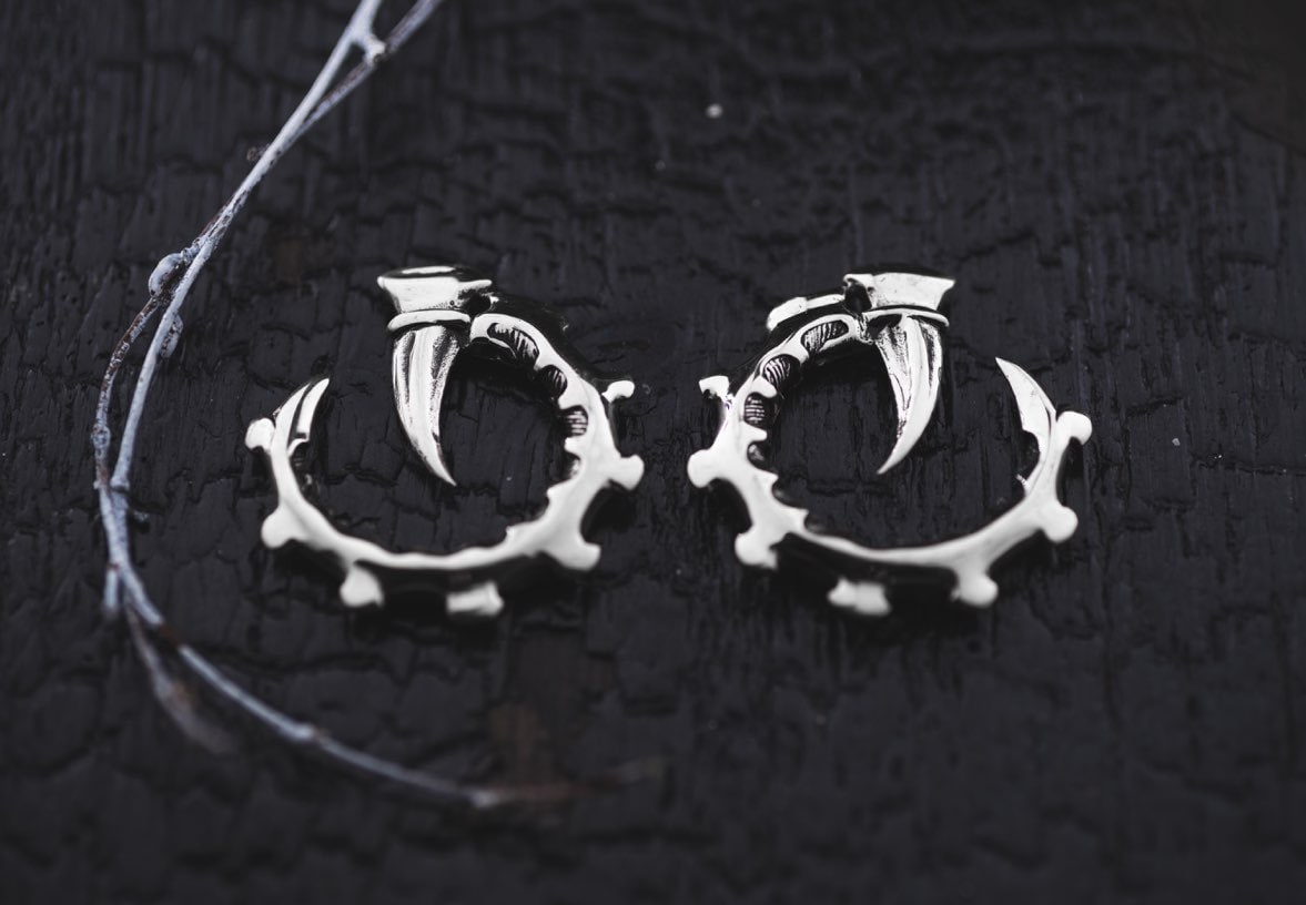 A pair of GIGER Biomechanical Open Ended Hoop Ear Weights in Silver by Kaya Movement, shaped like bear claws, rest on a textured black surface. A slender, dried twig lies beside them, adding a rustic, natural element to the composition. The earrings evoke post-apocalyptic body jewelry with intricate detailing on the claw and circular frame.