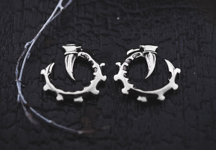 A pair of GIGER Biomechanical Open Ended Hoop Ear Weights in Silver by Kaya Movement, shaped like bear claws, rest on a textured black surface. A slender, dried twig lies beside them, adding a rustic, natural element to the composition. The earrings evoke post-apocalyptic body jewelry with intricate detailing on the claw and circular frame.
