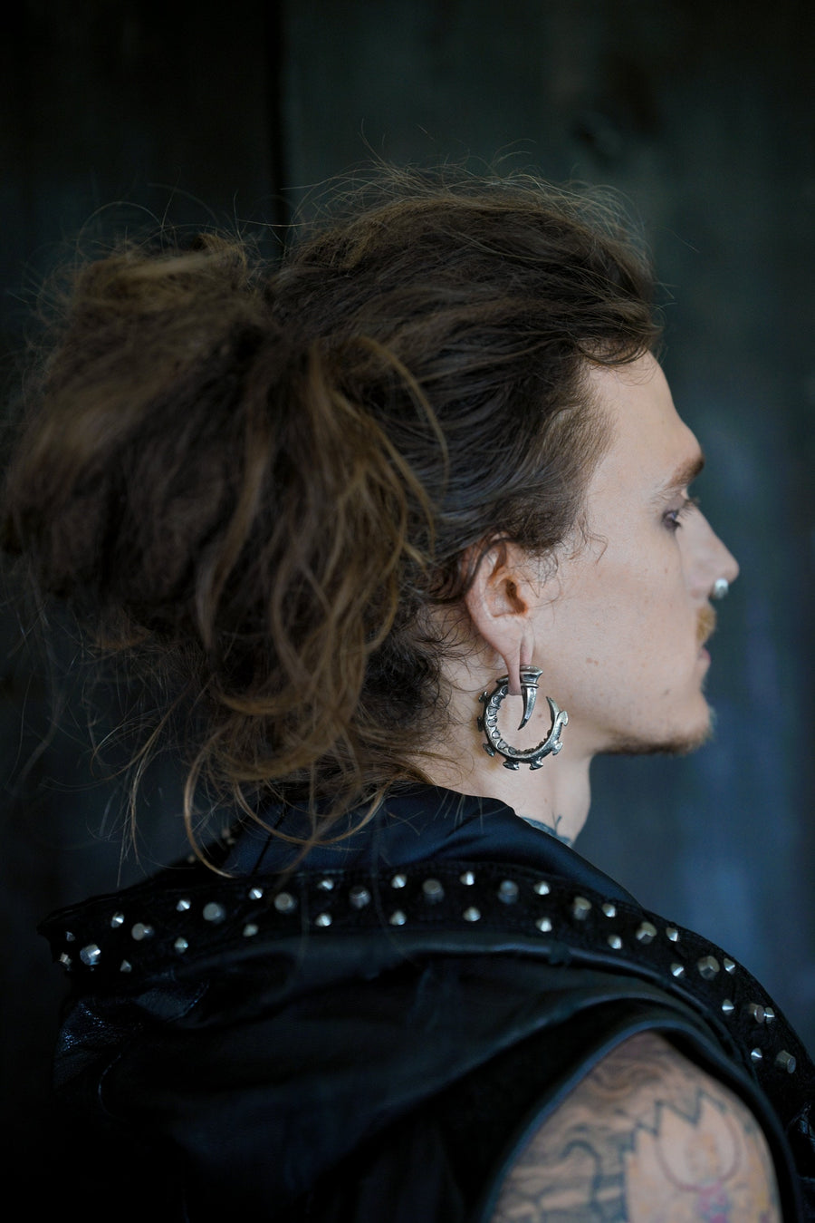 A person with long, messy brown hair tied back is shown in profile against a dark background. They are wearing a studded black leather garment and have elaborate gauge plug earrings and a nose ring, along with the GIGER Biomechanical Open Ended Hoop Ear Weights in Silver by Kaya Movement (0 gauge) and tattoos visible on their arm.