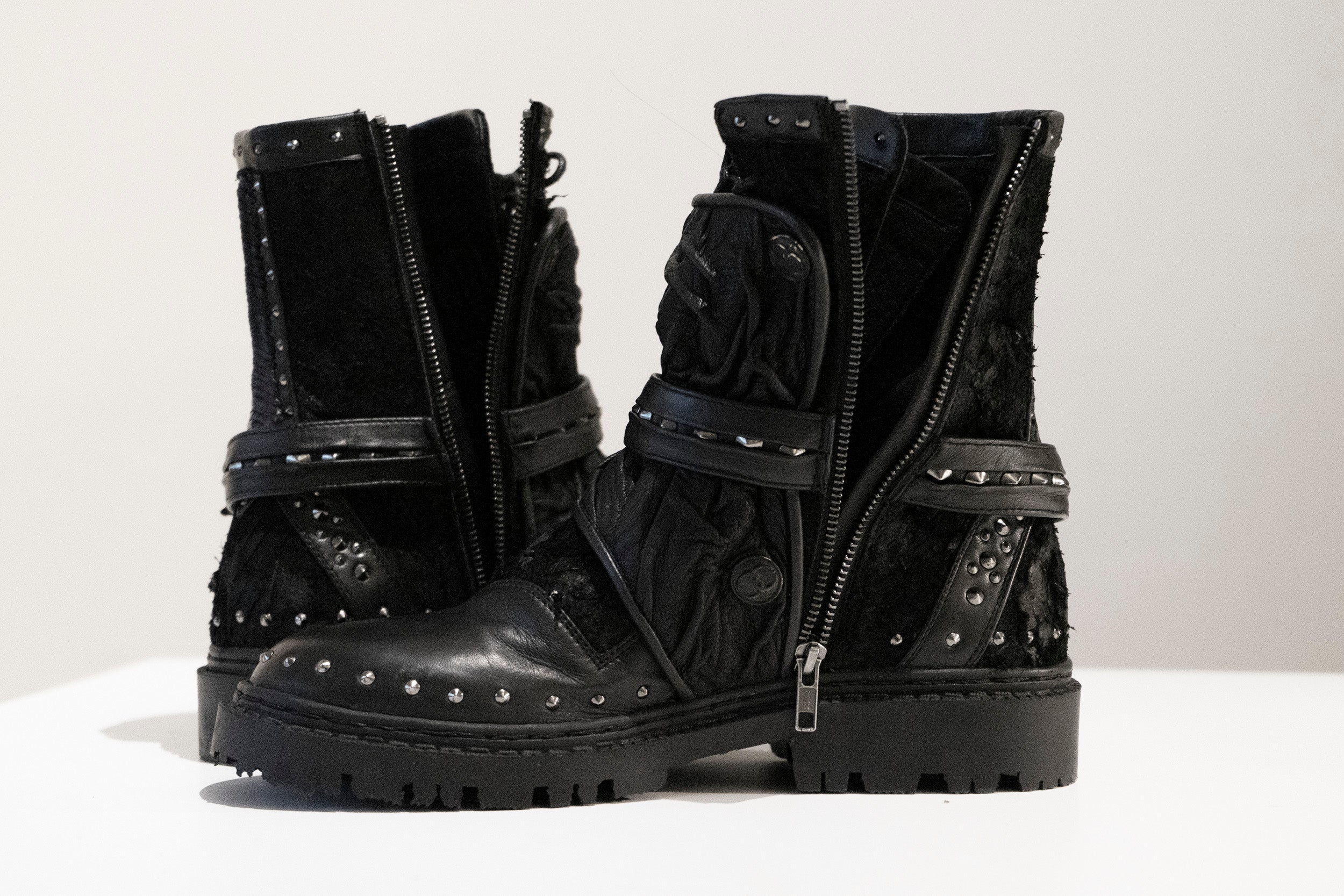 A pair of rugged design black, mid-calf leather boots featuring studded accents, chunky soles, and half-zipped fronts. Both boots have a textured finish and decorative straps with metal embellishments. These unisex MAGMA Wasteland Studded Leather Ankle Combat Boots by Kaya Movement are displayed on a white surface.