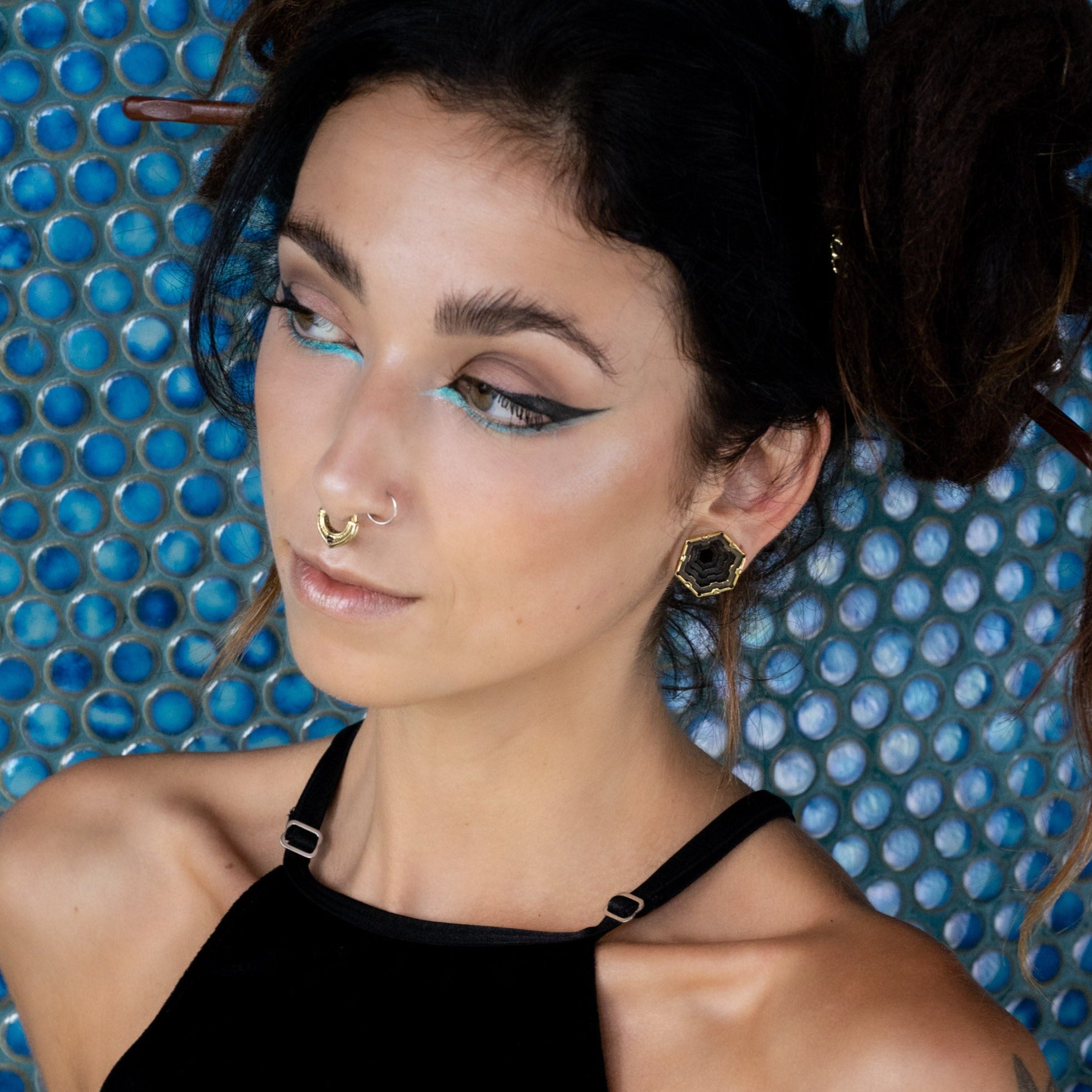 A woman with dark hair in an intricate braided updo stands against a textured, blue circular-patterned backdrop. She wears dramatic winged eyeliner, sky blue eyeshadow, a gold septum ring, HEXA Futuristic Geometry Hexagon Flesh Tunnels in Gold by Kaya Movement in her stretched ears, and a black strappy top while gazing to the side.