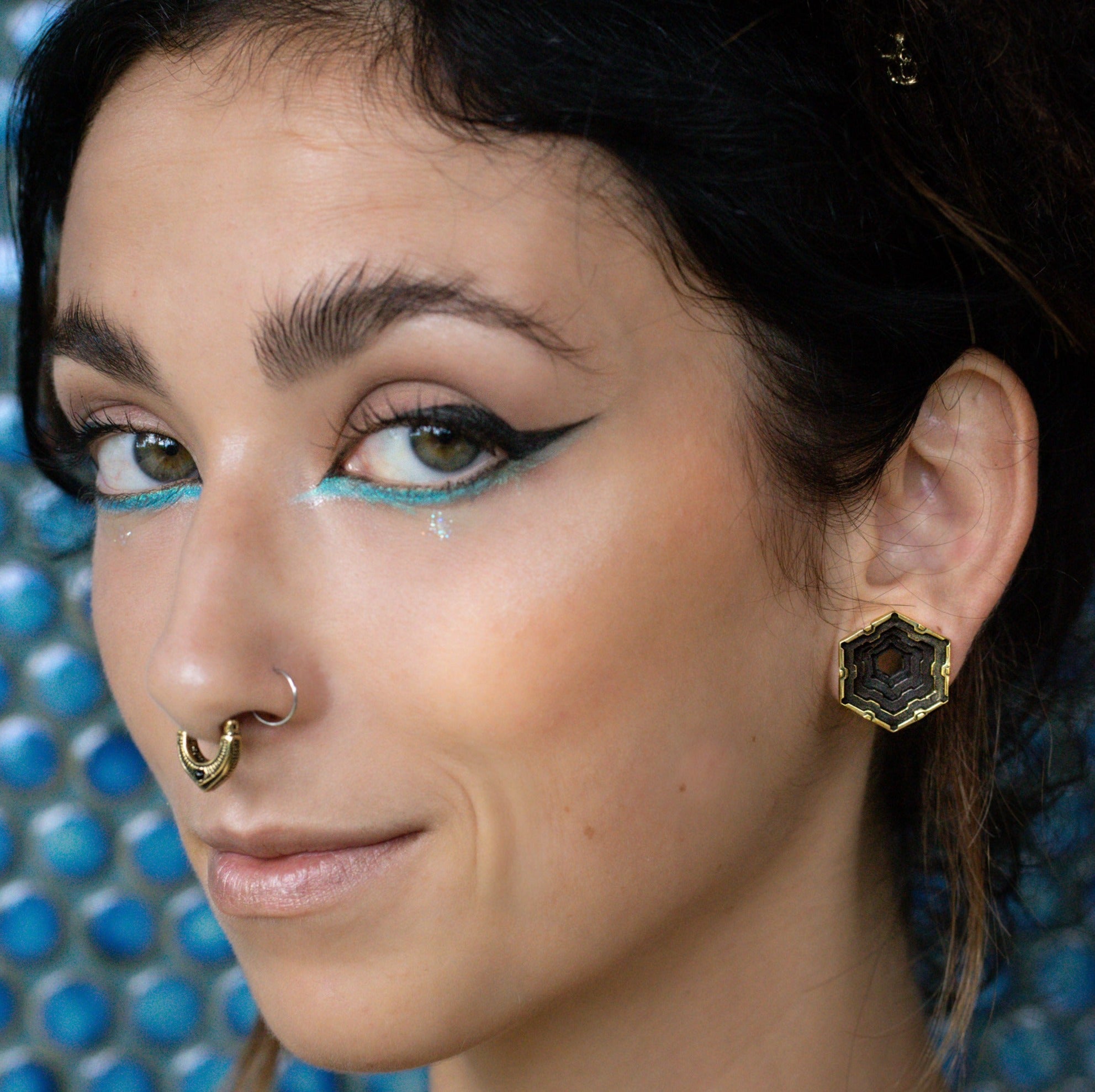 A person with dark hair and a nose ring is shown in a close-up. They are wearing HEXA Futuristic Geometry Hexagon Flesh Tunnels in Gold by Kaya Movement and have bold, winged eyeliner with a hint of blue eyeshadow beneath their eyes. The background features a blue and white circular pattern.