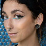 Close-up of a person with neatly groomed eyebrows and striking makeup, featuring thick winged eyeliner, aqua blue lower eyeliner, highlighter on the cheekbones, and a septum piercing. They are wearing HEXA Futuristic Geometry Hexagon Flesh Tunnels in Silver by Kaya Movement. The background has a blue dotted pattern.