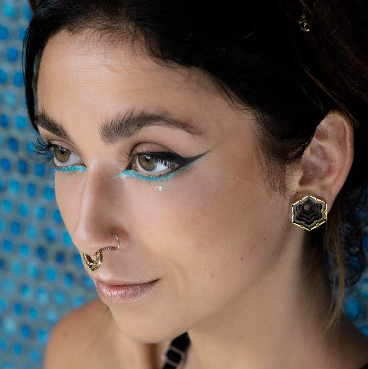 A woman with dark hair, adorned in a gold septum ring, is shown in close-up, gazing to the side. She has striking eye makeup featuring blue and black eyeliner and is wearing HEXA Futuristic Geometry Hexagon Flesh Tunnels in Gold by Kaya Movement in her stretched ears. The background showcases a blue patterned design.