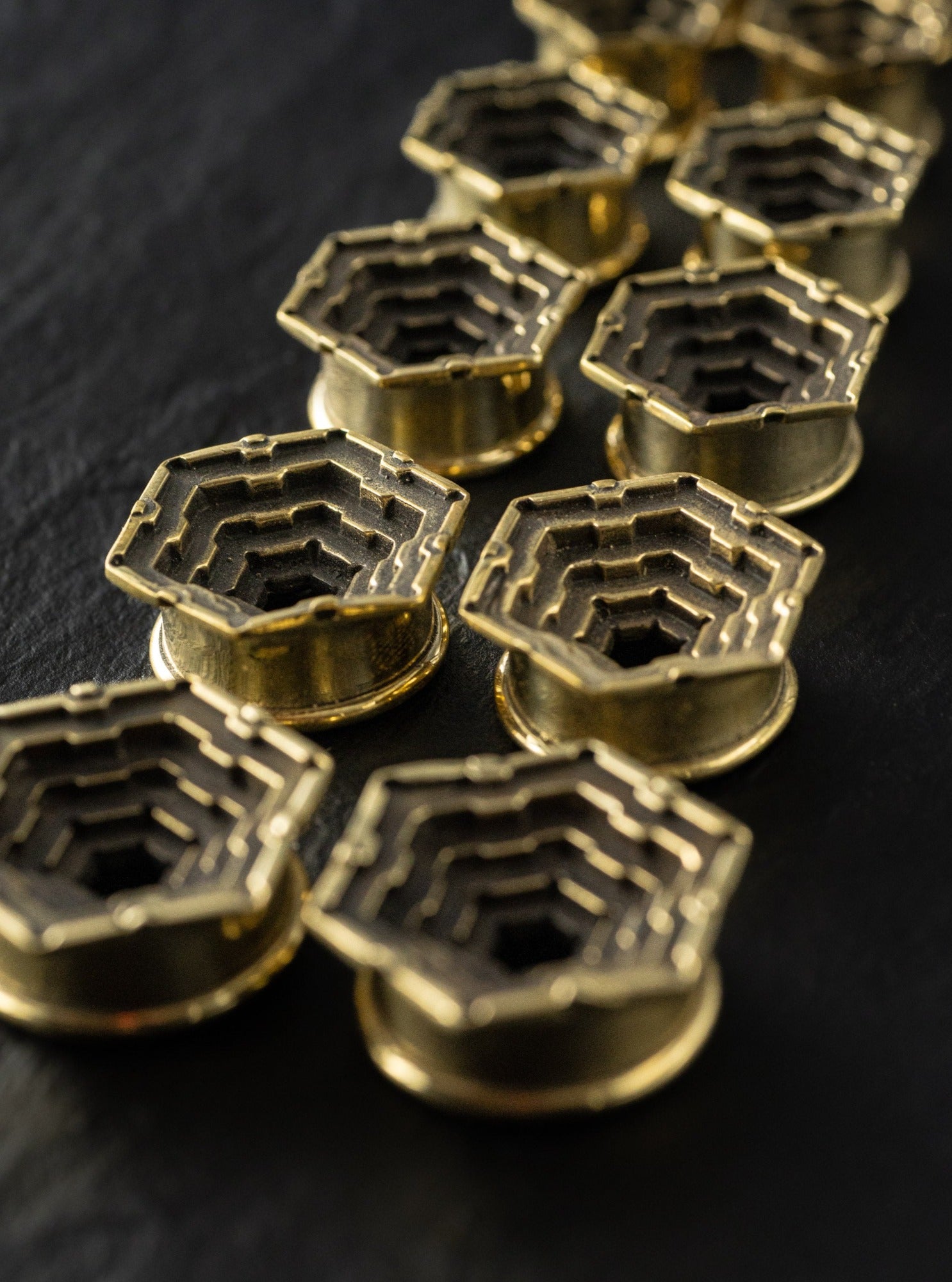 A close-up of multiple intricate, HEXA Futuristic Geometry Hexagon Flesh Tunnels in Gold by Kaya Movement, lined up diagonally on a dark background. The components have layers creating a stepped, three-dimensional effect.