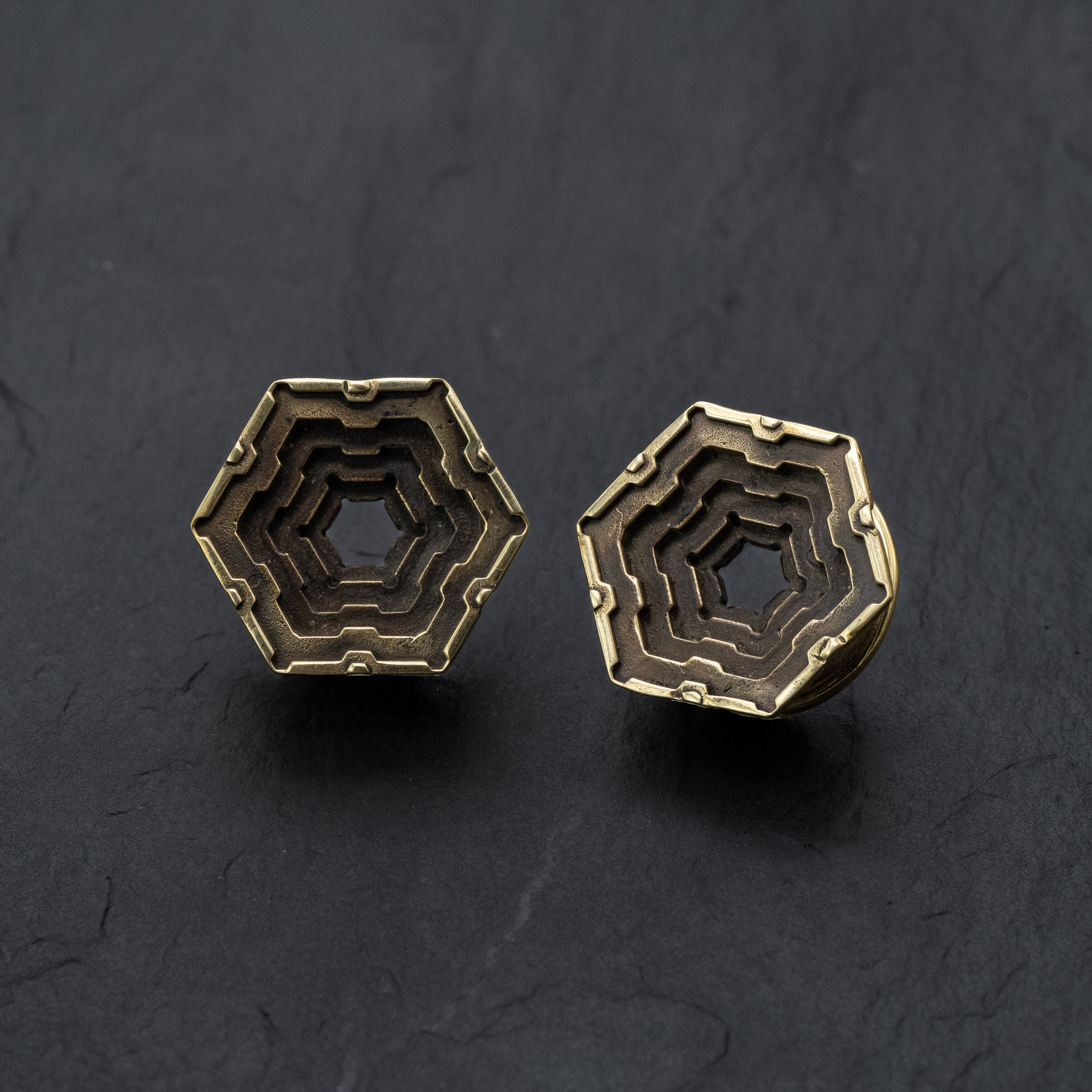 A striking pair of HEXA Futuristic Geometry Hexagon Flesh Tunnels in Gold by Kaya Movement, available in sizes from 12mm to 24mm. These elegant pieces are evocative of visionary AI plug earrings and boast a layered, stepped geometric pattern within the hexagon, creating a captivating three-dimensional effect. The photograph showcases them against a dark textured background.