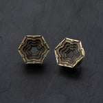 A striking pair of HEXA Futuristic Geometry Hexagon Flesh Tunnels in Gold by Kaya Movement, available in sizes from 12mm to 24mm. These elegant pieces are evocative of visionary AI plug earrings and boast a layered, stepped geometric pattern within the hexagon, creating a captivating three-dimensional effect. The photograph showcases them against a dark textured background.