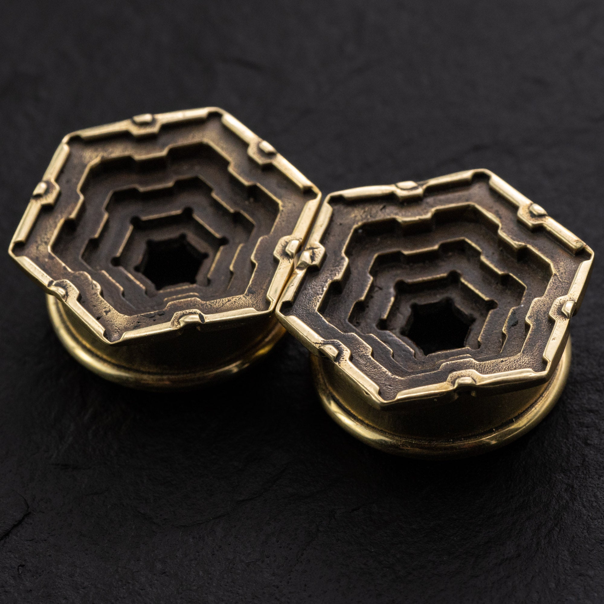 A close-up image of a pair of HEXA Futuristic Geometry Hexagon Flesh Tunnels in Gold, by Kaya Movement. The multi-layered geometric designs within the hexagonal shapes showcase intricate patterns, reminiscent of visionary AI plug earrings. Set against a dark background, the image highlights the metallic texture and craftsmanship of these 12mm to 24mm pieces.