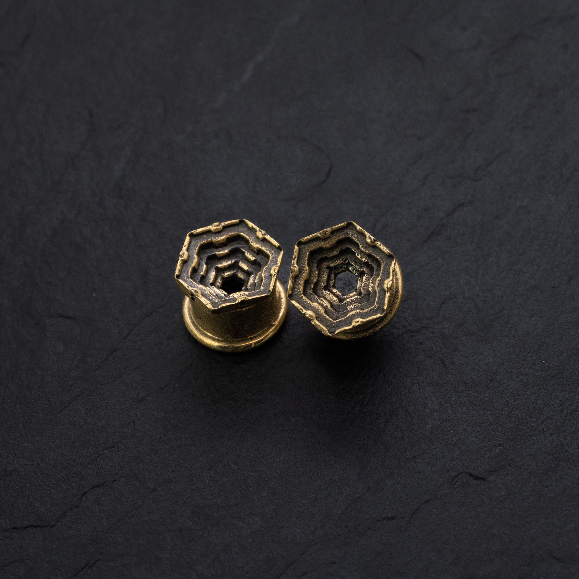 A pair of HEXA Futuristic Geometry Hexagon Flesh Tunnels in Gold from Kaya Movement, featuring an intricate hexagonal, layered maze-like design set against a dark, textured backdrop. The multilayered pattern adds depth and complexity to these accessories, reminiscent of visionary AI-inspired plug earrings for stretched ears. Available in sizes ranging from 12mm to 24mm.