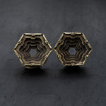 A pair of hexagon-shaped metal earrings with an intricate layered design and a dark patina is set against a black background to highlight their detailed craftsmanship and geometric structure, reminiscent of the HEXA Futuristic Geometry Hexagon Flesh Tunnels in Gold by Kaya Movement, available in sizes from 12mm to 24mm.