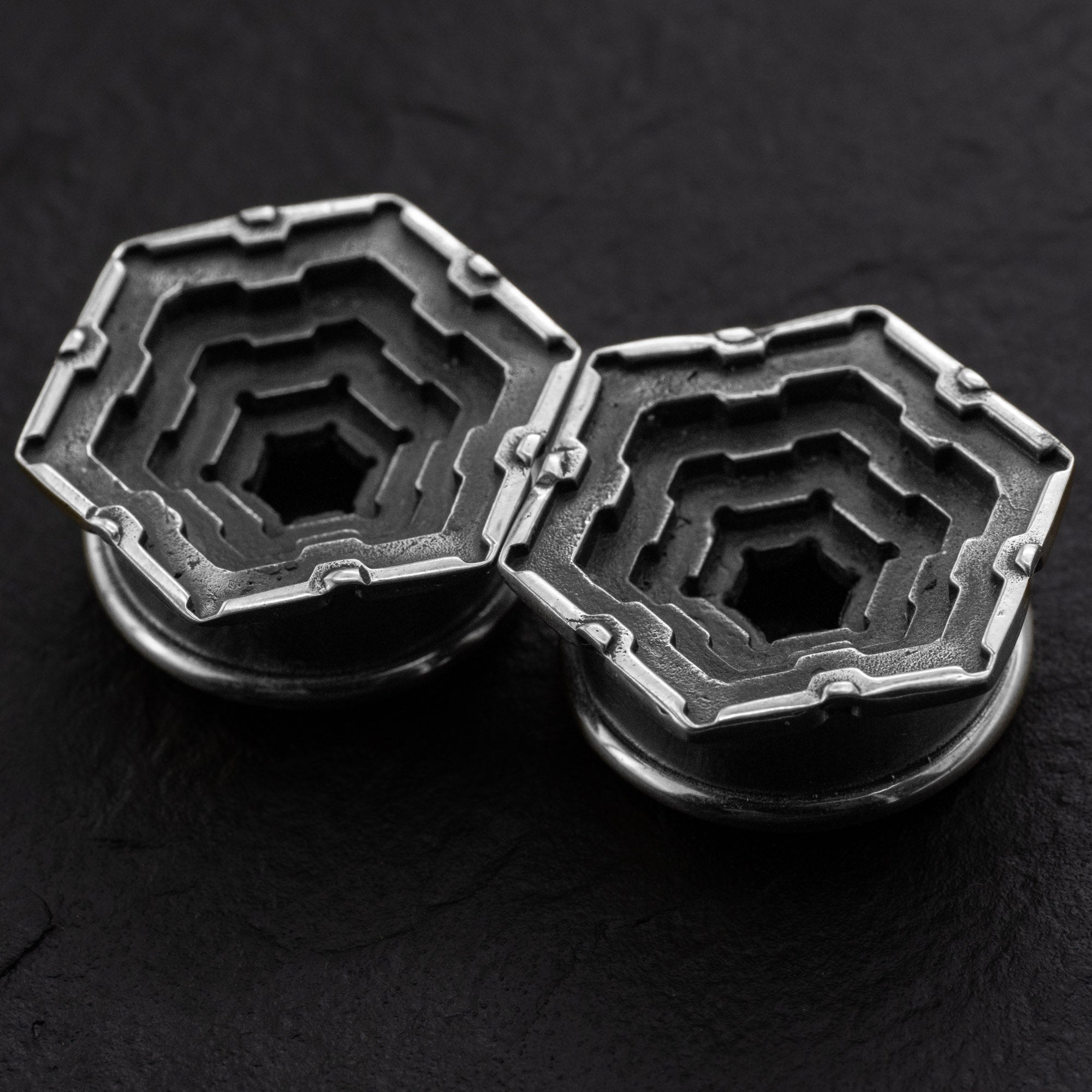 A close-up of two Kaya Movement HEXA Futuristic Geometry Hexagon Flesh Tunnels in silver, featuring intricate, stepped designs that resemble nested hexagons. These precision-crafted objects, reminiscent of body jewelry like flesh tunnel earrings for stretched ears, range in sizes from 12mm to 24mm and are placed side by side on a dark, textured surface. Their symmetrical design showcases high attention to detail.