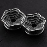 A close-up of two Kaya Movement HEXA Futuristic Geometry Hexagon Flesh Tunnels in silver, featuring intricate, stepped designs that resemble nested hexagons. These precision-crafted objects, reminiscent of body jewelry like flesh tunnel earrings for stretched ears, range in sizes from 12mm to 24mm and are placed side by side on a dark, textured surface. Their symmetrical design showcases high attention to detail.