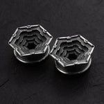 Close-up of the HEXA Futuristic Geometry Hexagon Flesh Tunnels by Kaya Movement in silver, featuring an industrial-style hexagonal design. The flesh tunnels exhibit a layered, gear-like pattern reminiscent of body jewelry like cufflinks and are set against a dark, textured background. Available in sizes ranging from 12mm to 24mm.