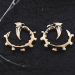 Close-up shot of GIGER Biomechanical Open Ended Hoop Ear Weights in Gold, designed by Kaya Movement to resemble animal claws gripping circular, gear-like objects. Perfect for dystopian festivals, the unisex 0 gauge plug earrings are laid out on a dark textured surface.