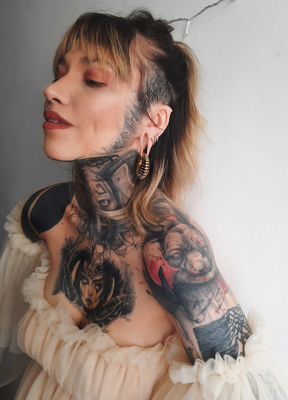A person with a nose ring and multiple tattoos on their neck and shoulder, including a detailed portrait, smiles while looking up. They wear large hoop earrings and a ruffled off-the-shoulder top accessorized with Kaya Movement's TRILOBITE Large Biomechanical Clicker Ear Weights in Gold Brass (0 gauge). The background is a plain light-colored wall.