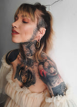 A person with a nose ring and multiple tattoos on their neck and shoulder, including a detailed portrait, smiles while looking up. They wear large hoop earrings and a ruffled off-the-shoulder top accessorized with Kaya Movement's TRILOBITE Large Biomechanical Clicker Ear Weights in Gold Brass (0 gauge). The background is a plain light-colored wall.