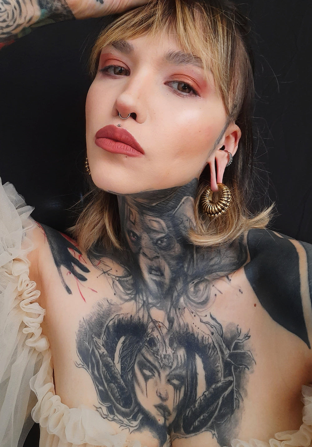 A person with blonde hair, red eyeshadow, and dark lipstick poses against a dark background. They have multiple tattoos, including a prominent one of a horned figure on their chest. Adorned in post-apocalyptic body jewelry and wearing the TRILOBITE Large Biomechanical Clicker Ear Weights in Gold Brass by Kaya Movement in 0 gauge, they also have tribal gauge earrings, gold earrings, and a nose ring while wearing a ruffle off-the-shoulder top.
