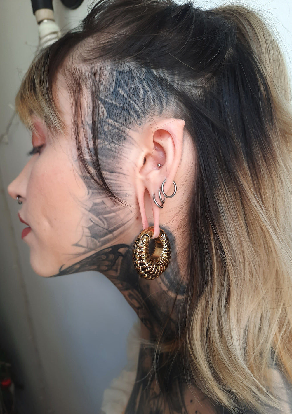 A person with intricate black tattoos on their neck and side of their face is shown in profile, exuding a post-apocalyptic body jewelry vibe. They have multiple piercings on their ear, including Kaya Movement's TRILOBITE Large Biomechanical Clicker Ear Weights in Gold Brass (0 gauge), smaller hoop earrings, and a flesh tunnel. Their hair is partially shaved and dyed.
