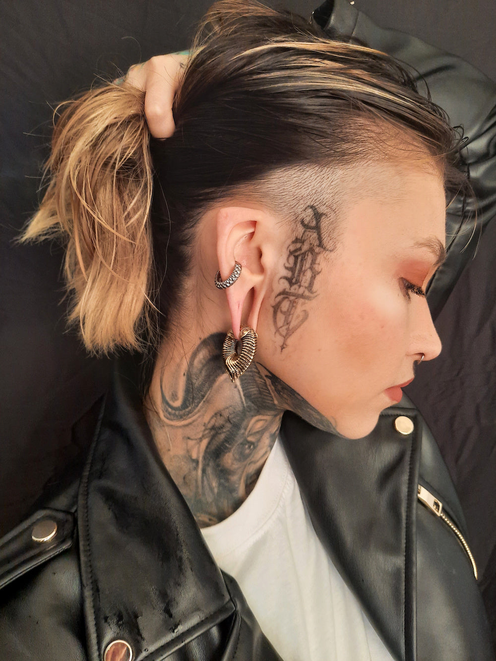 A person with a side-swept haircut and shaved side is seen in profile, showcasing numerous tattoos on their neck and scalp, as well as multiple piercings featuring MARVDASHT Medium Cyborg Clicker Ear Weights in Gold (3/18" gauge) by Kaya Movement. The person is wearing a black leather jacket and a white shirt.