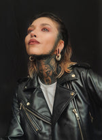 A person with short, partially dyed hair looks upward against a dark background. They have tattoos on their neck and chin, piercings including a nose ring, and are wearing MARVDASHT Medium Cyborg Clicker Ear Weights in Gold by Kaya Movement. Clad in a leather jacket zipped halfway over a white shirt, they exude an edgy style.