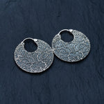 A pair of Kaya Movement's KONI Tribal Shipibo Endless Hoop Disc Earrings in Silver (18 gauge) are placed on a dark, textured surface. These 925 sterling silver tribal body jewelry pieces are detailed with intricate geometric patterns and textures, featuring repeating lines and shapes that give them a sophisticated and artistic appearance.