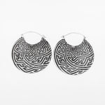 Introducing the SAYAGATA Oversized Tribal Disc Hoop Earrings by Kaya Movement. These 16-gauge, circular earrings in silver feature intricate, maze-like patterns with detailed geometric designs that create a textured appearance. Perfect for a boho statement, they are displayed against a plain white background.