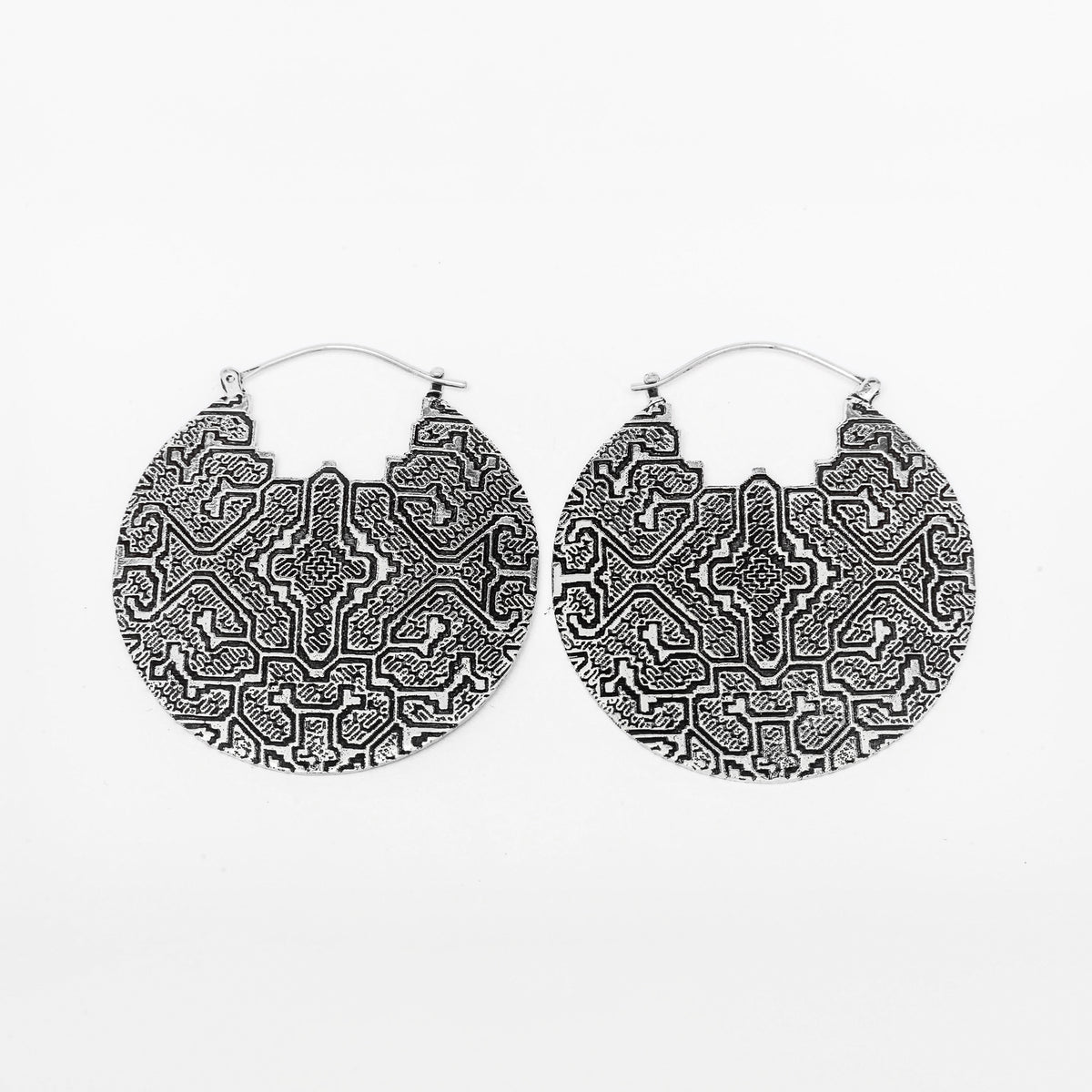 The PANCHA Oversized Tribal Shipibo Disc Earrings in Silver by Kaya Movement feature a pair of large, circular statement earrings with intricate, symmetrical geometric patterns etched into their surfaces. These 16-gauge silver earrings have a hook clasp and are beautifully displayed against a white background.