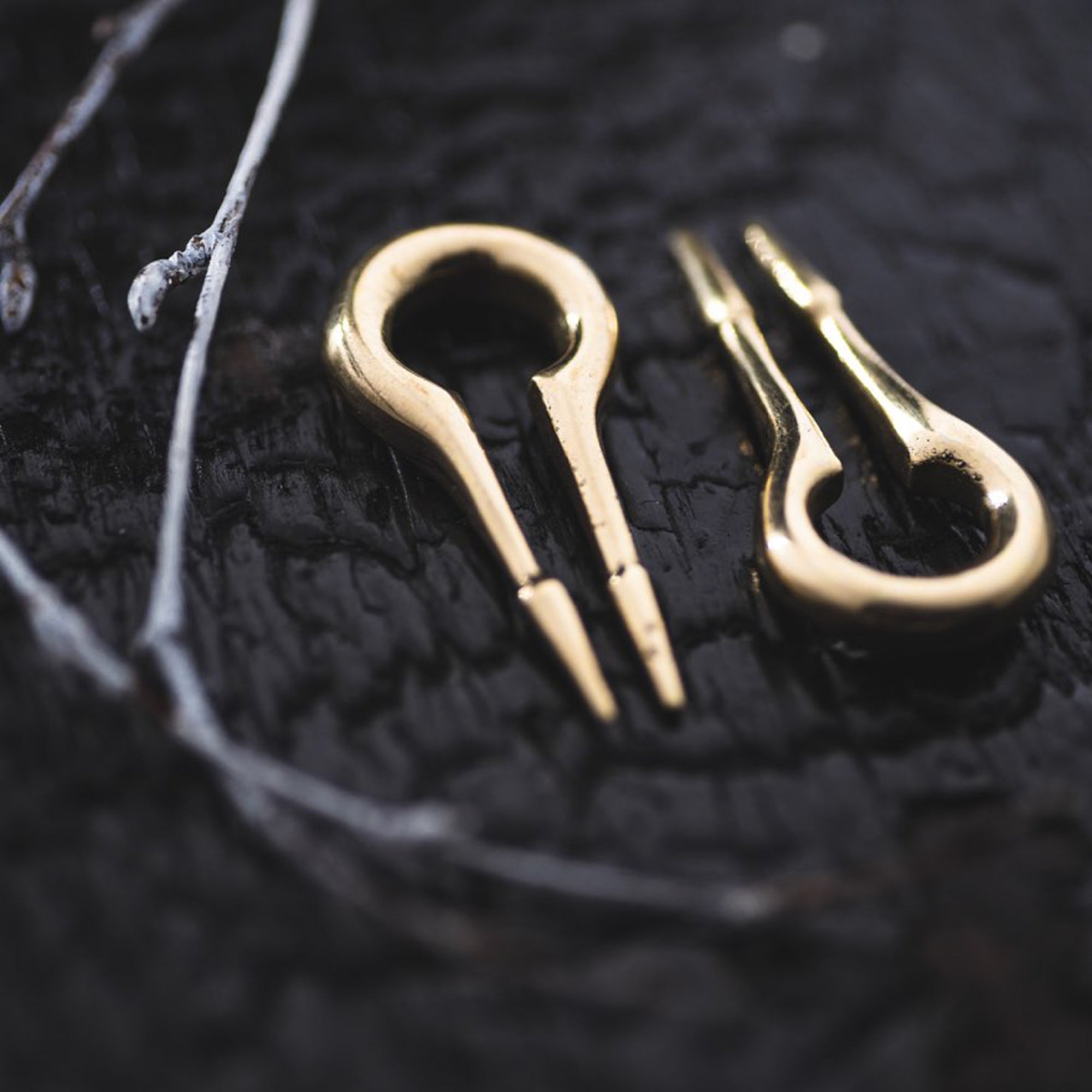 Close-up of two CALYPSO Minimalist Keyhole Ear Weights in Gold by Kaya Movement, lying on a textured dark surface. Resembling delicate musical instruments, one ear weight is face-up while the other is side-up, near a small twig. The image captures the intricate details and reflections of these 2-gauge ear weights.