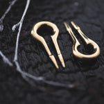 Close-up of two CALYPSO Minimalist Keyhole Ear Weights in Gold by Kaya Movement, lying on a textured dark surface. Resembling delicate musical instruments, one ear weight is face-up while the other is side-up, near a small twig. The image captures the intricate details and reflections of these 2-gauge ear weights.