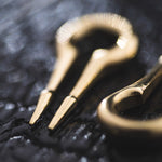 Close-up of a brass jew's harp, an ancient musical instrument, placed on a dark, textured surface. The image highlights the instrument's intricate design and polished finish, reminiscent of Kaya Movement's CALYPSO Minimalist Keyhole Ear Weights in Gold | 2 gauge, emphasizing its reflective quality and fine craftsmanship.