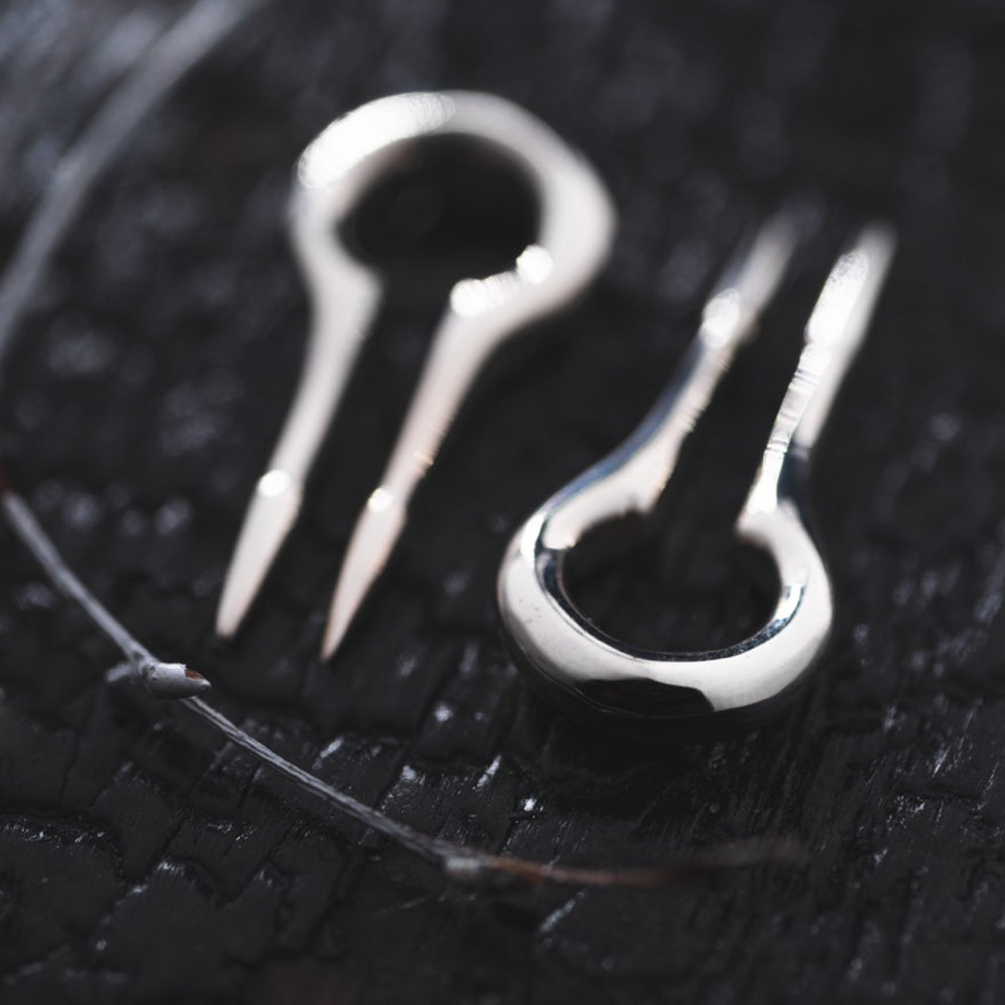 Close-up of two CALYPSO Minimalist Keyhole Ear Weights in 925 Silver by Kaya Movement resting on a dark textured surface. One ear weight has its prongs positioned close together, while the other has a small gap between its prongs. A thin string with small knots, akin to that used in Kaya Movement's 925 silver earrings, is visible next to the ear weights.
