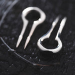 Close-up of two CALYPSO Minimalist Keyhole Ear Weights in 925 Silver by Kaya Movement resting on a dark textured surface. One ear weight has its prongs positioned close together, while the other has a small gap between its prongs. A thin string with small knots, akin to that used in Kaya Movement's 925 silver earrings, is visible next to the ear weights.