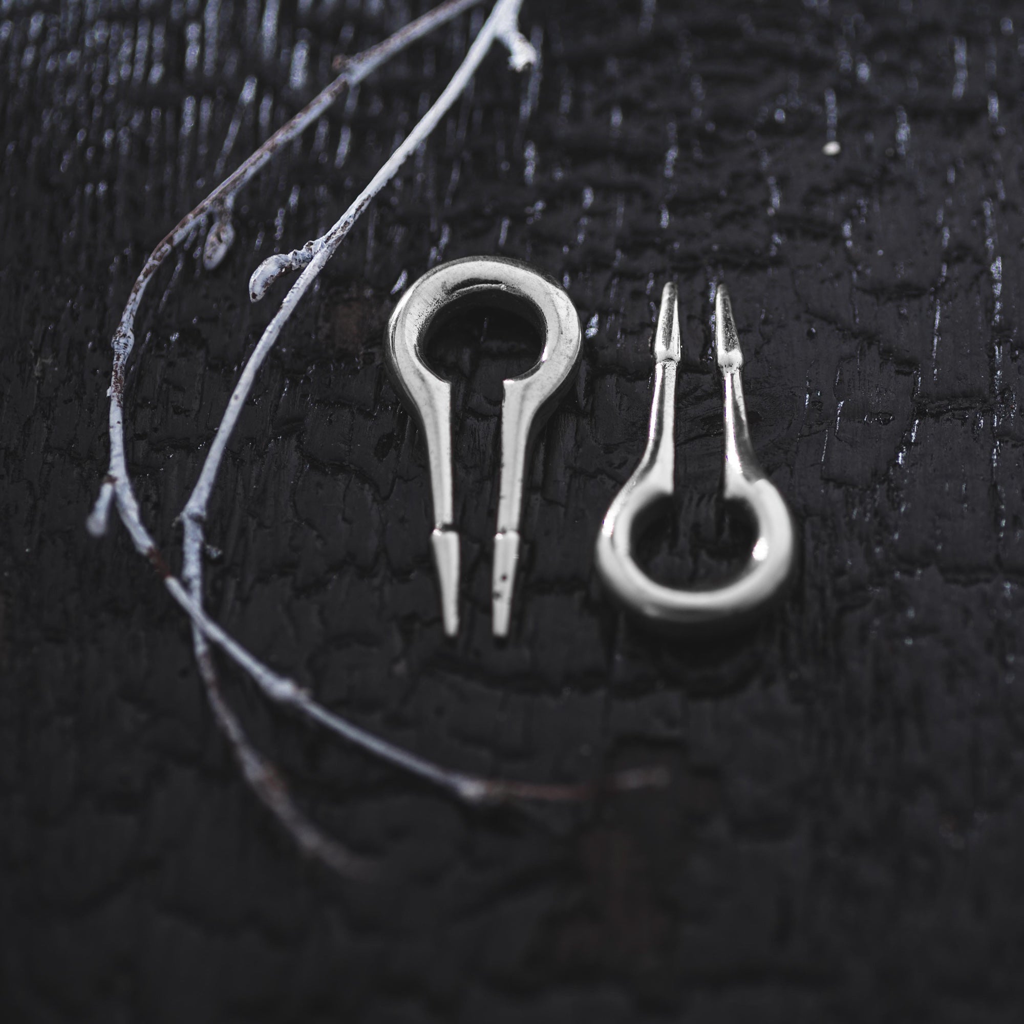 A close-up of two small metal tools with forked ends placed on a dark, textured background. To the left of the tools, thin white branches rest on the surface, adding contrast to the scene. The setting appears moody and detailed, reminiscent of displays for Kaya Movement's CALYPSO Minimalist Keyhole Ear Weights in 925 Silver, 2 gauge.